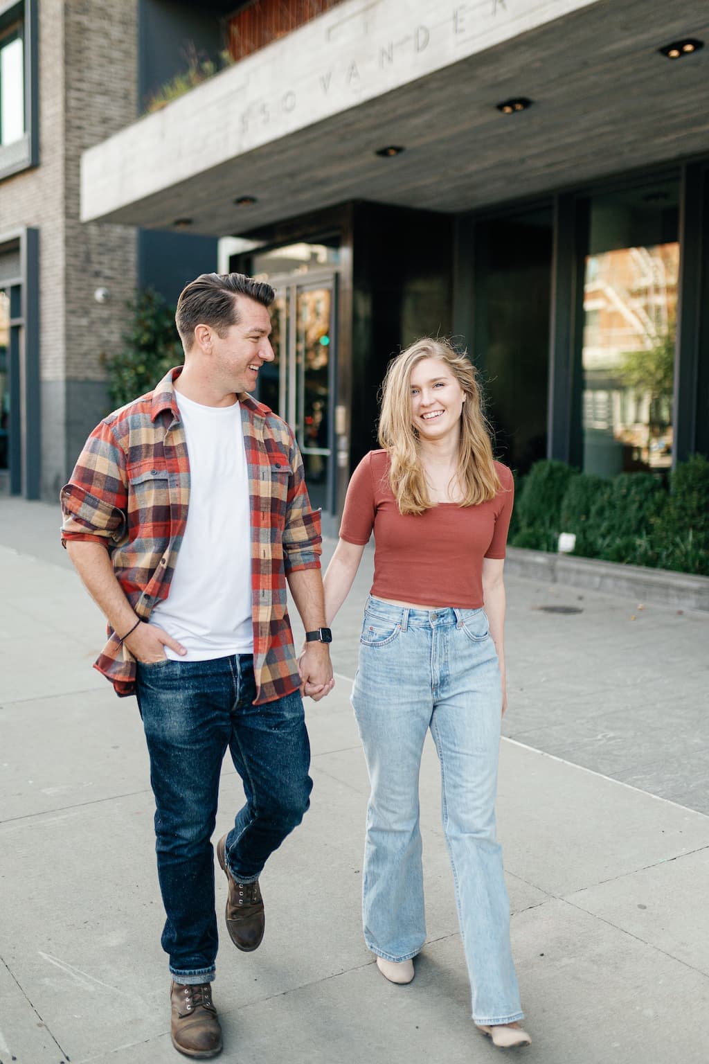 Eli and Georgia, in front of a Brooklyn apartment building, holding hands and walking. Eli is looking at Georgia, and Georgia is looking at the camera.